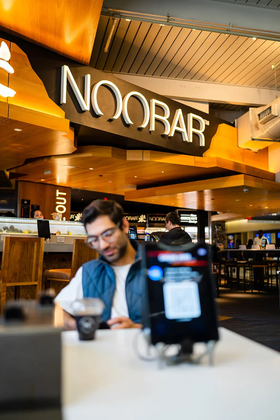 Person using a smartphone in a modern interior setting with 'NOOBAR' sign in background
