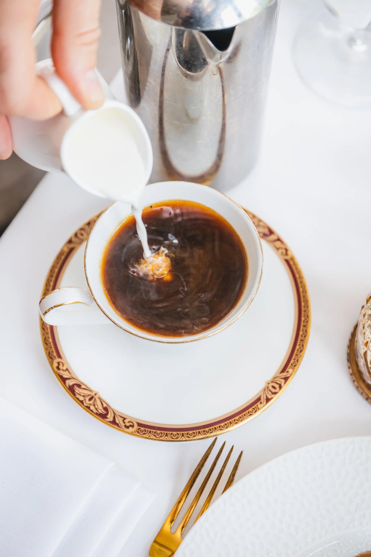 Person pouring milk into a cup of Bean2Bean coffee on a decorative plate with a gold rim.