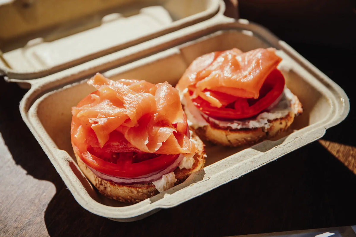 Two sandwiches with salmon and tomatoes in a cardboard container on a wooden surface.