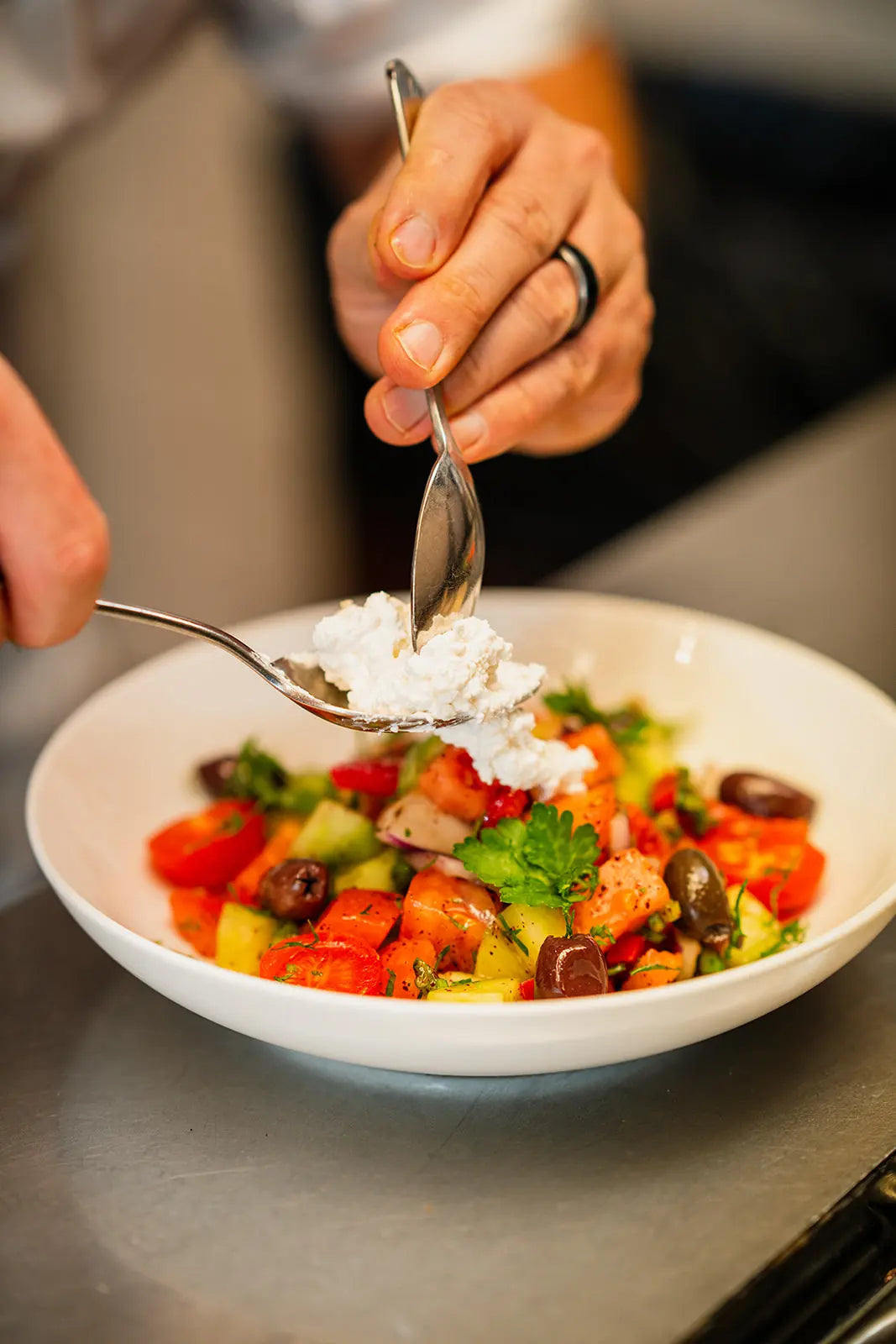 Person adding a spoonful of white sauce to a colorful salad in a white bowl.