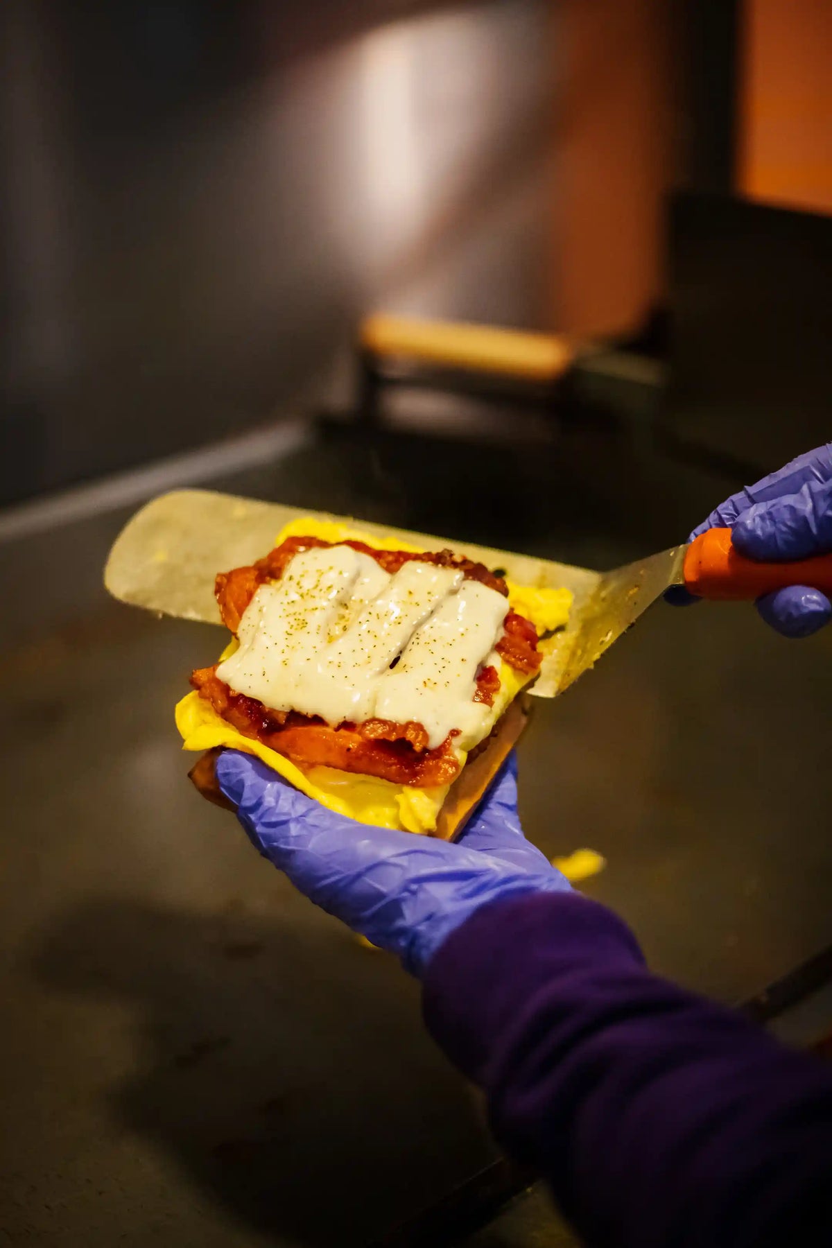 Hand with blue glove holding a sandwich with egg, cheese, and bacon, using scissors on a dark surface.
