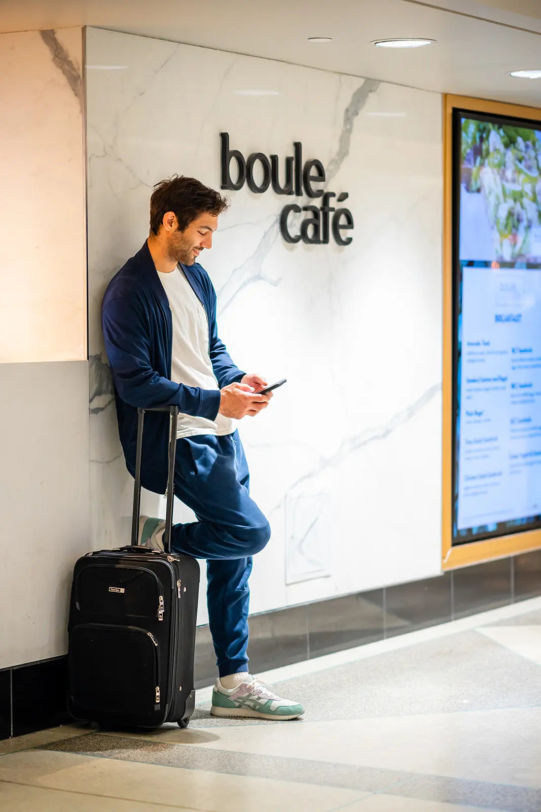 Man with a suitcase leaning against a wall with 'boule cafe' signage.