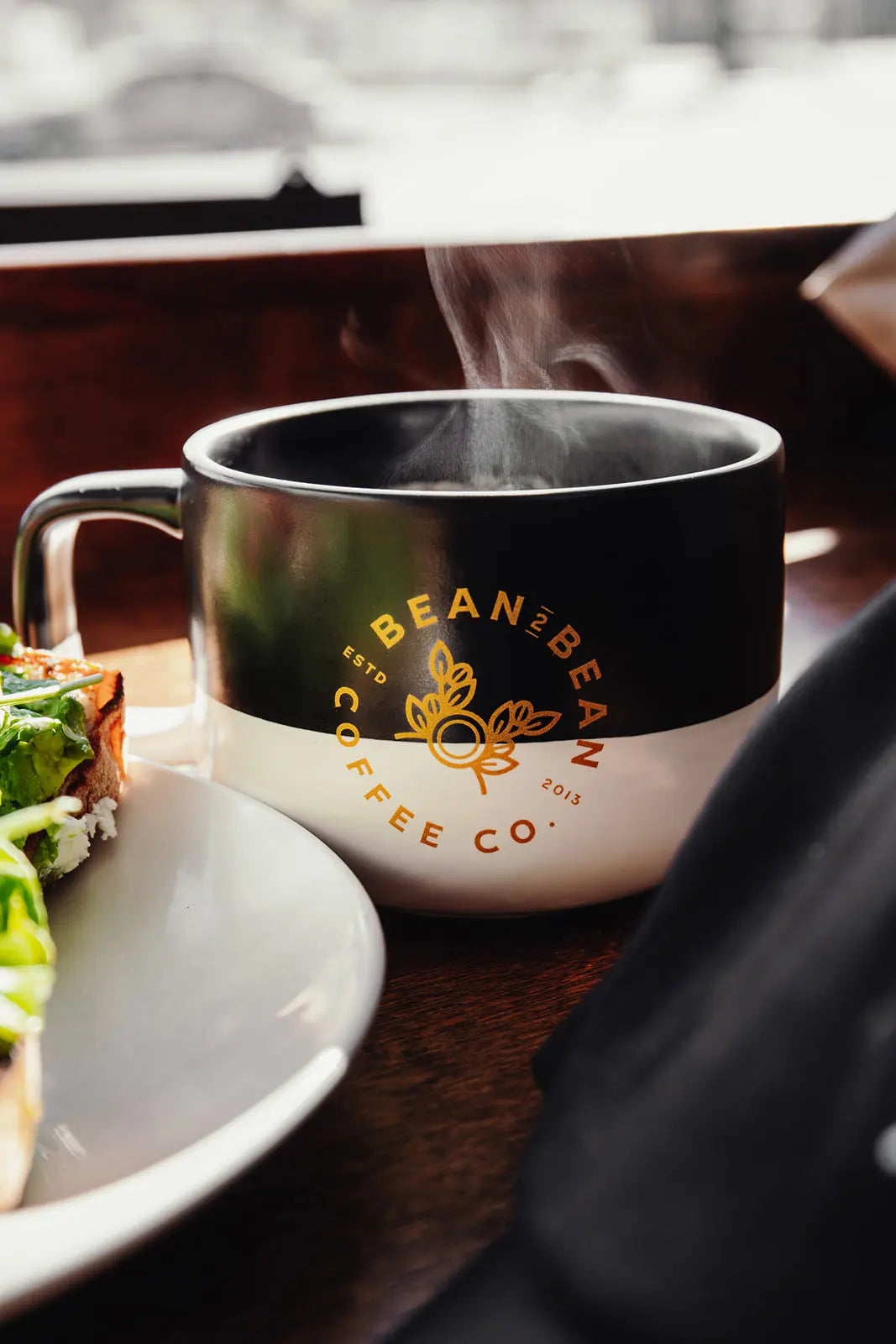 Steaming coffee cup from Bean2Bean Coffee Co. on a table with food.