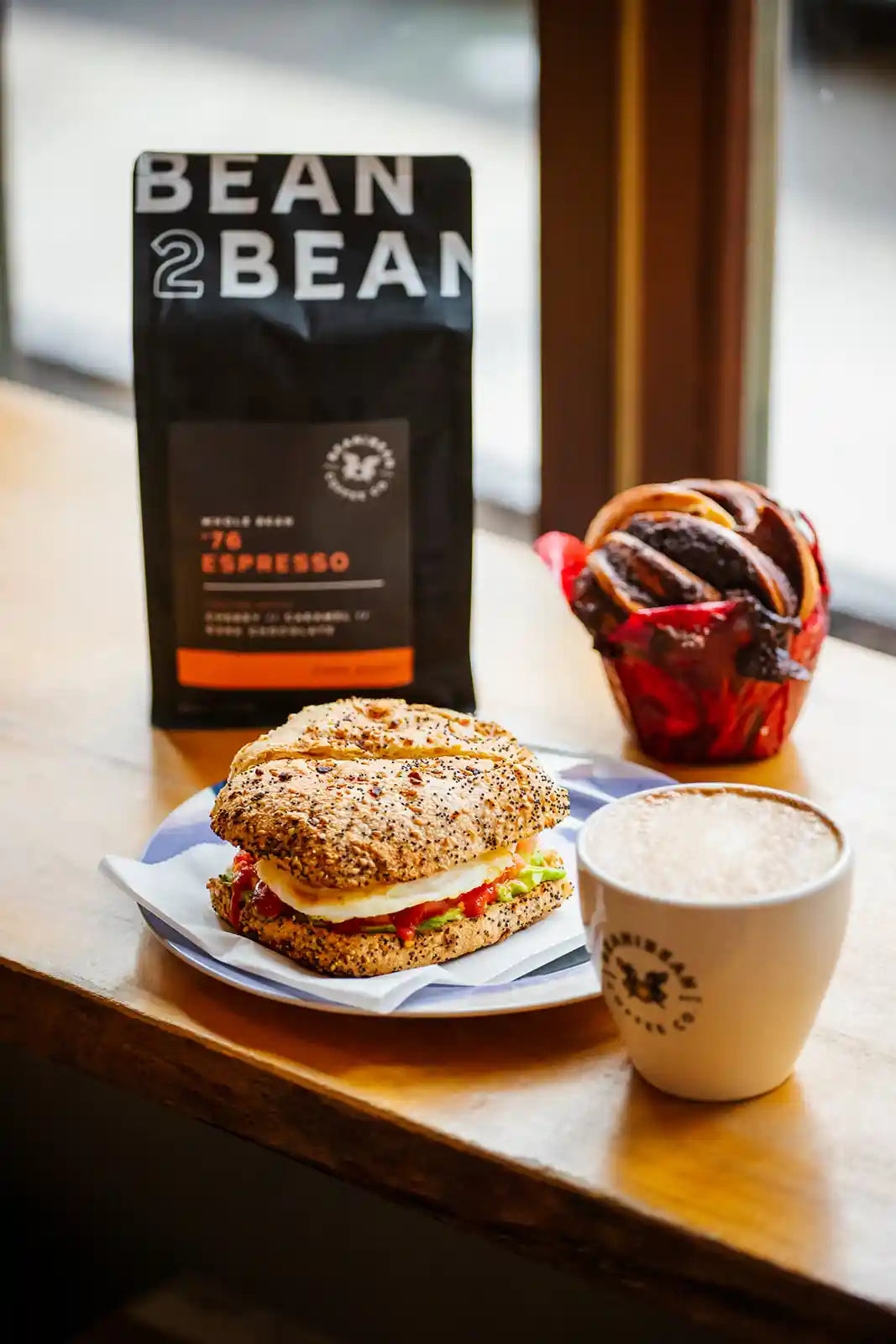 Sandwich, coffee, and 'Bean2Bean' coffee package on a wooden table.