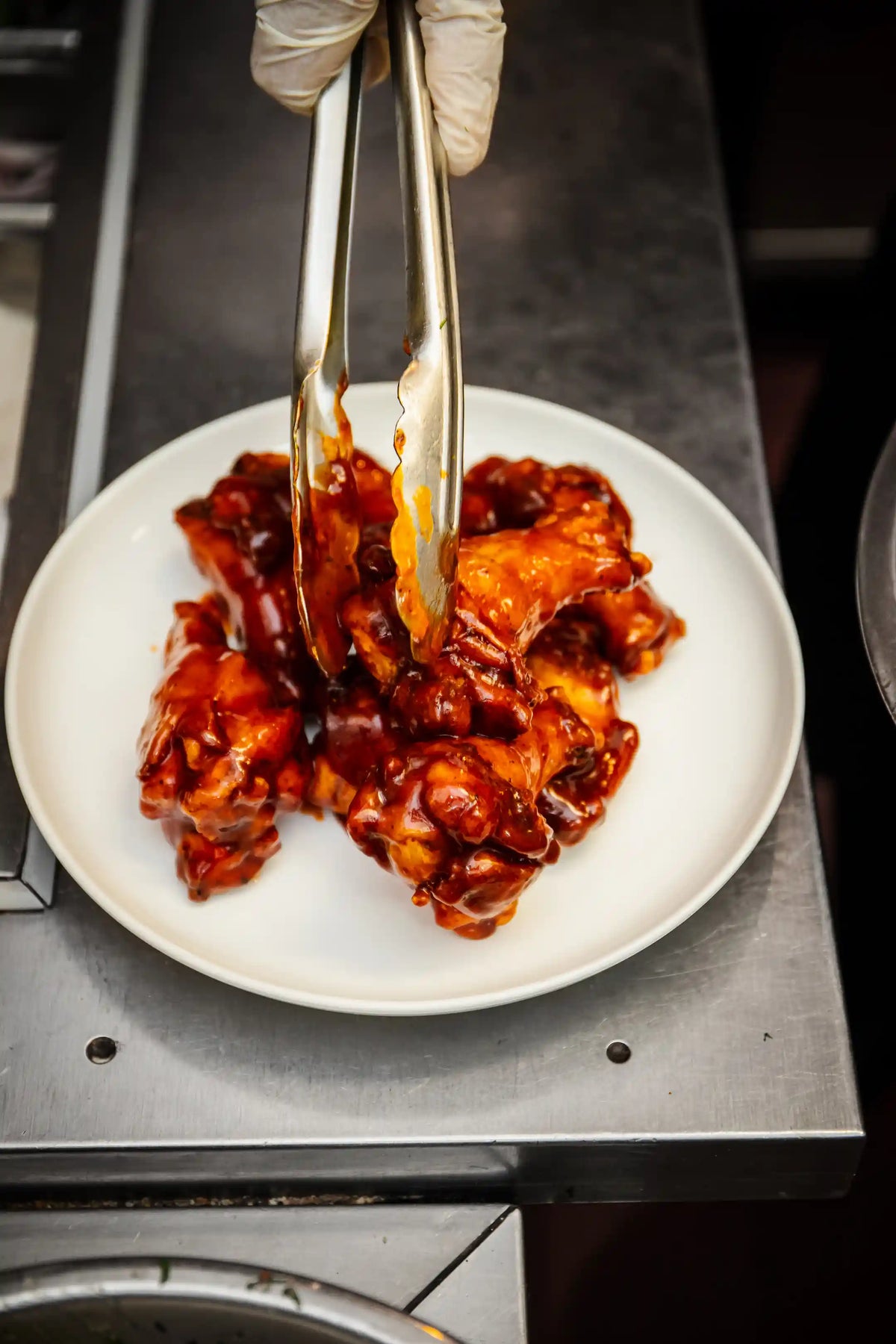 Sauced hot wings being served on a white plate with tongs, on a metallic surface.