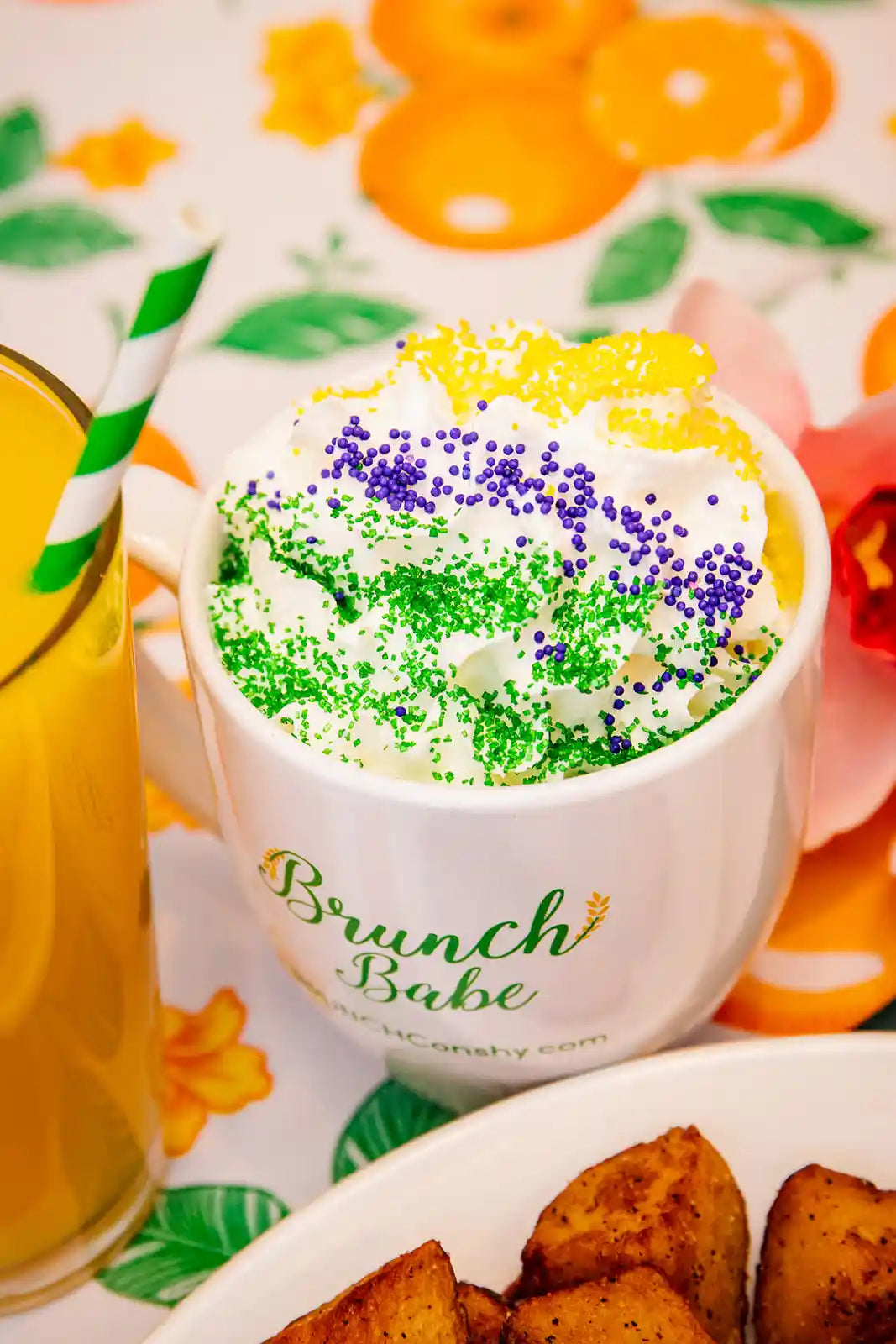 Cup of coffee with colorful sprinkles, glass of orange juice, and plate of food on a floral tablecloth.