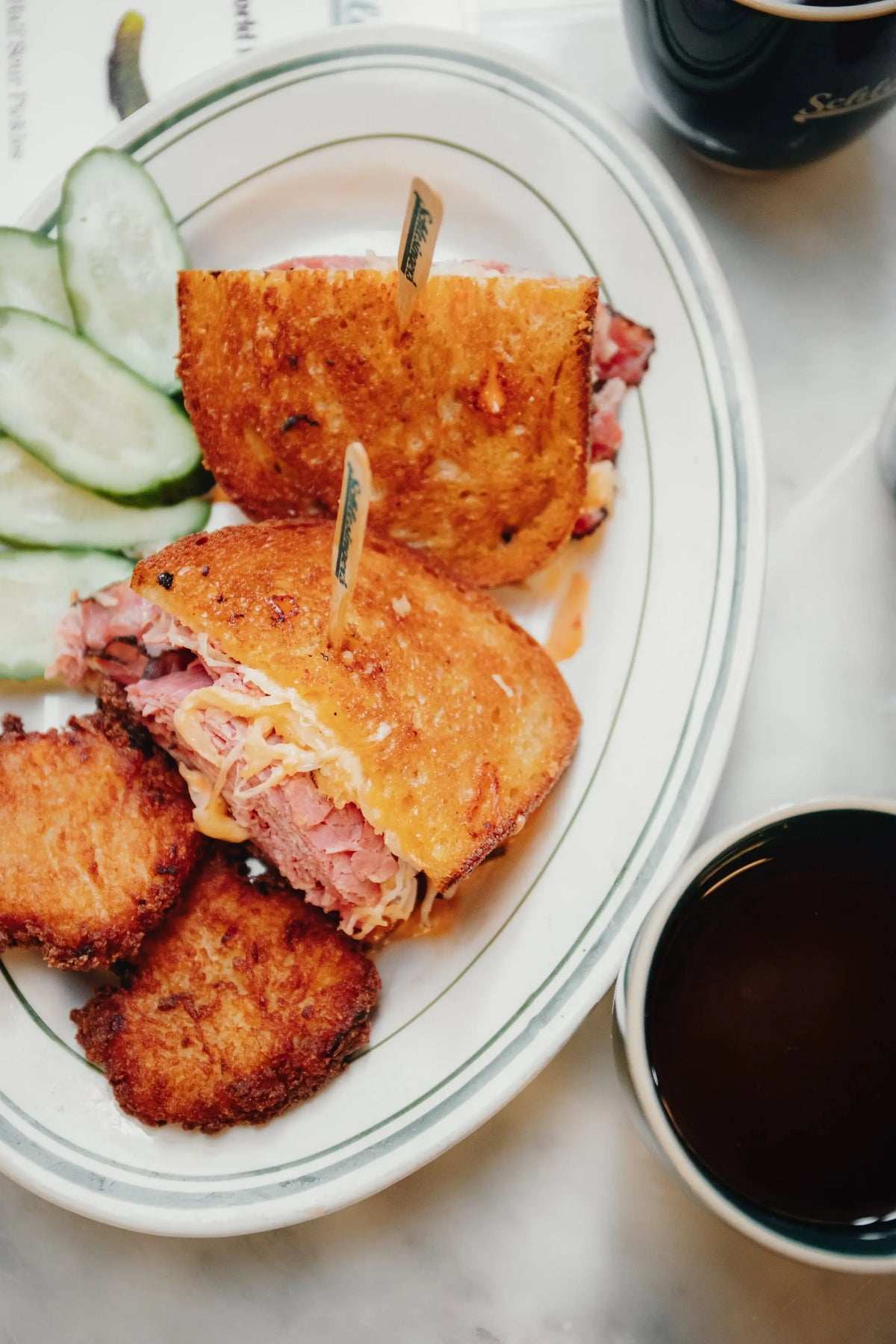 Fried sandwich cut in half on a plate with cucumbers, accompanied by two cups of coffee.