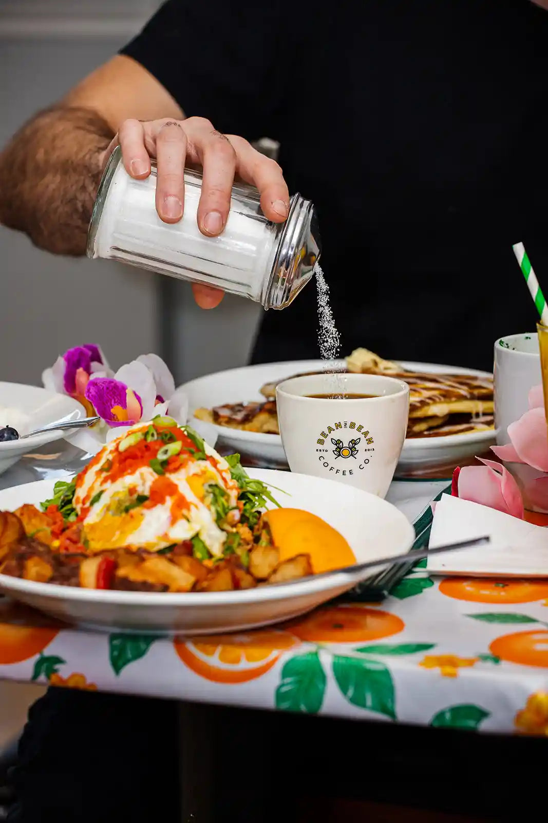 Person pouring sugar from a shaker into their coffee at a dining table with pancakes.