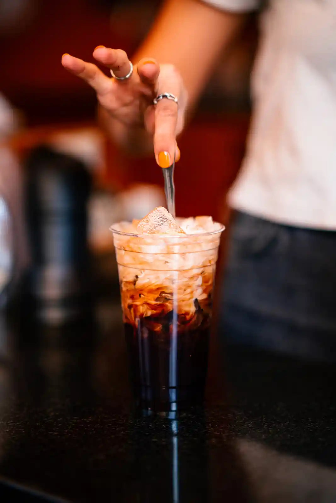 Person stirring an iced coffee drink with a spoon in a glass on a bar counter