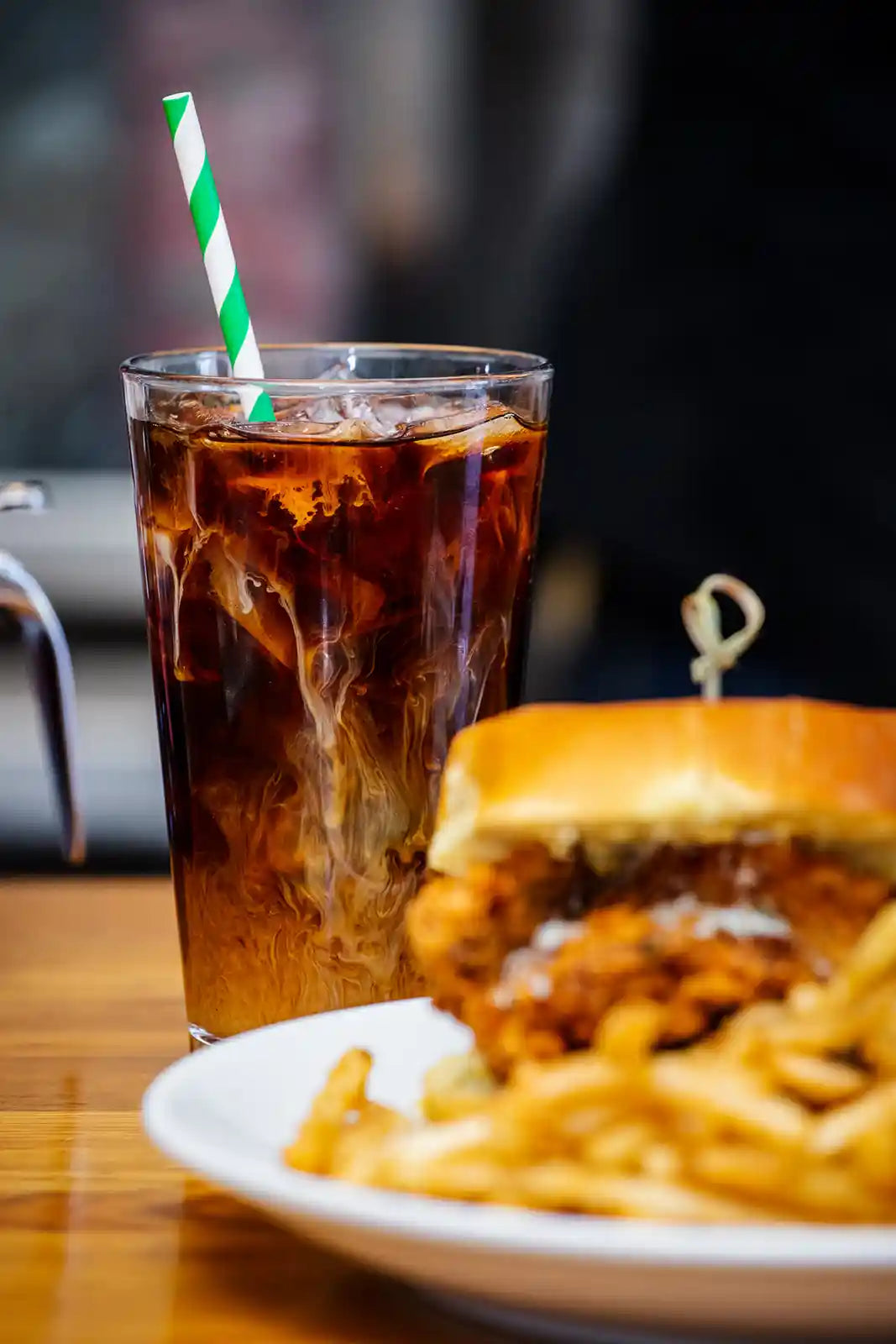 Glass of iced coffee with a straw next to a burger and fries on a wooden table.
