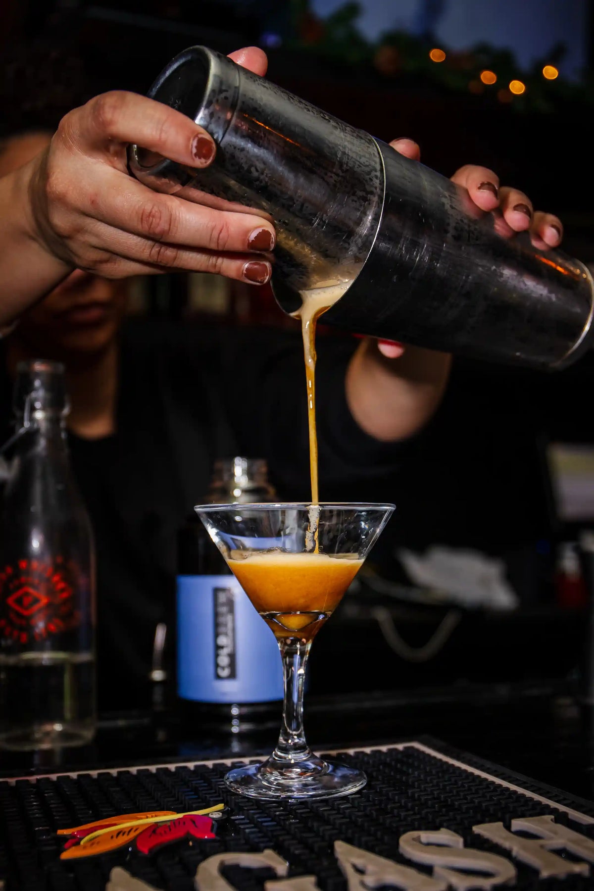 Person pouring an espresso martini cocktail from a shaker into a glass on a bar counter.
