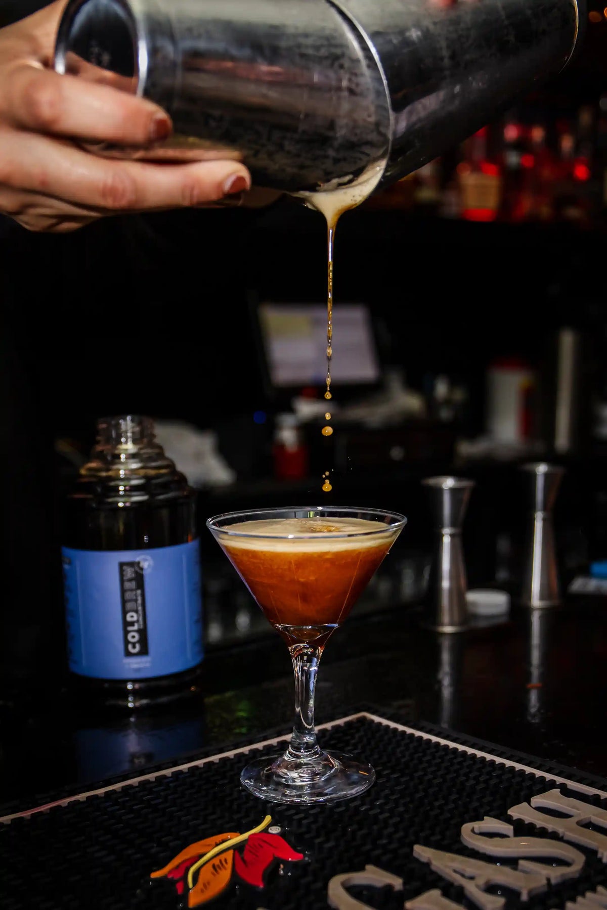 Person pouring an espresso martini cocktail from a shaker into a glass on a bar counter. Bean2Bean cold brew bottle in the background