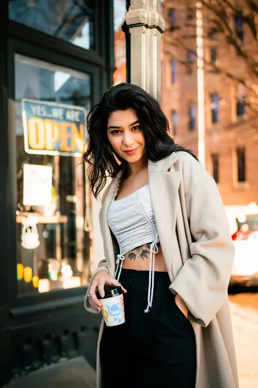 Woman standing on a city street holding a drink, with a building and 'Open' sign in the background.