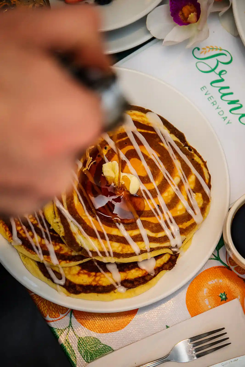 Person drizzling syrup over a stack of spiral batter pancakes on a plate with a decorative tablecloth.