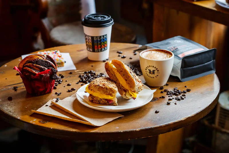 Wooden table with a sandwich, coffee, and muffin in a cozy setting