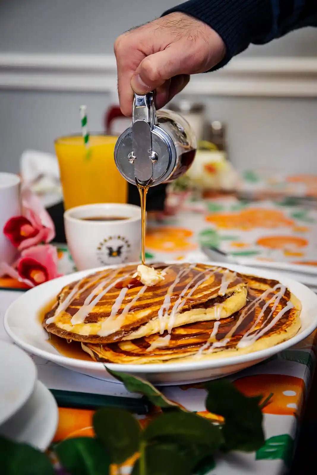 Person pouring syrup onto pancakes on a plate with a cup of coffee and orange juice in the background.