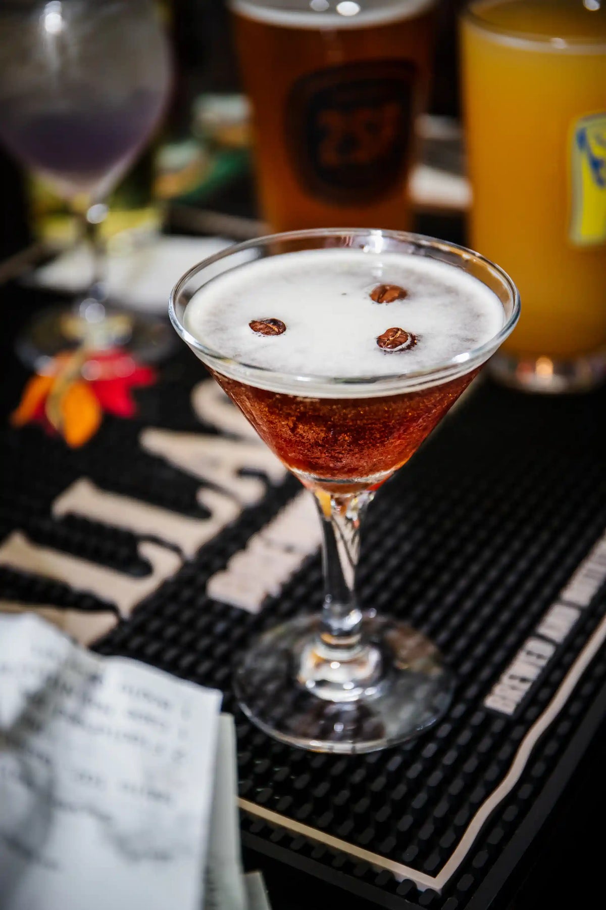 Espresso martini topped with coffee beans in a martini glass on a bar mat with other drinks in the background.