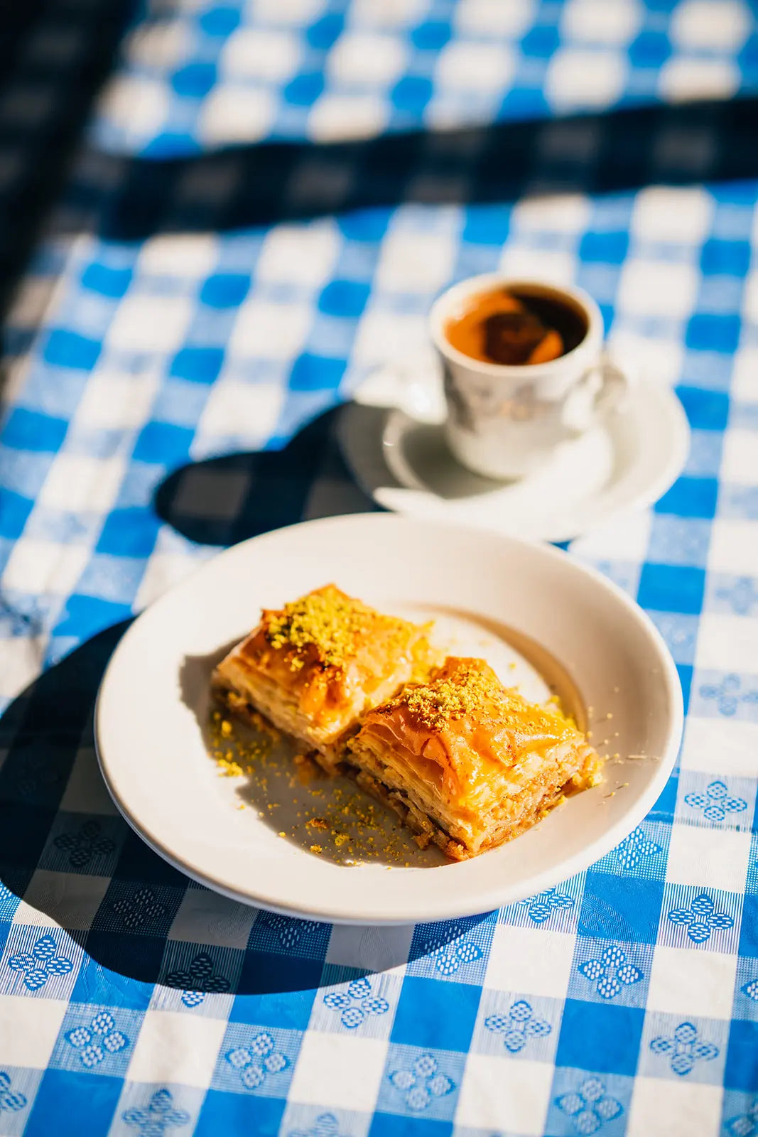 two slices of baklava on a white plate with a cup of Bean2Bean coffee on a blue and white checkered tablecloth.