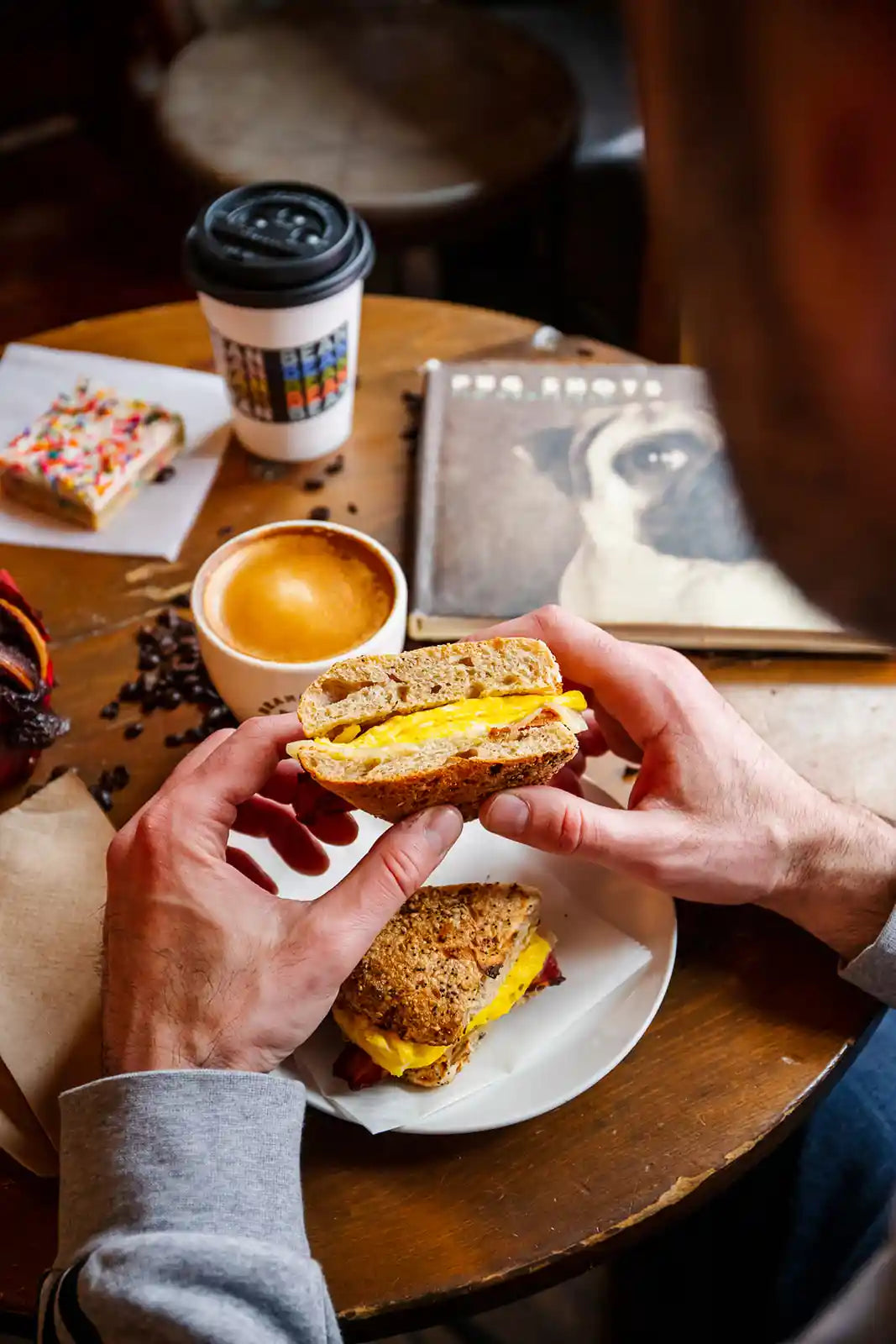 Person holding a sandwich with a cup of Bean2Bean coffee on a wooden table
