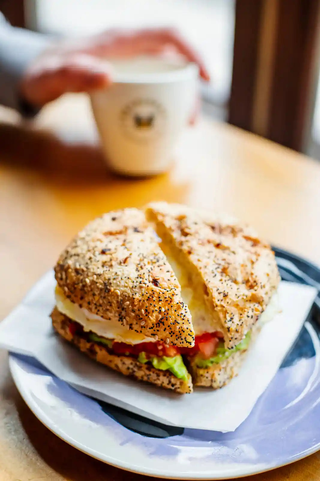 Bagel sandwich on a plate with a blurred background of a person holding a coffee cup.