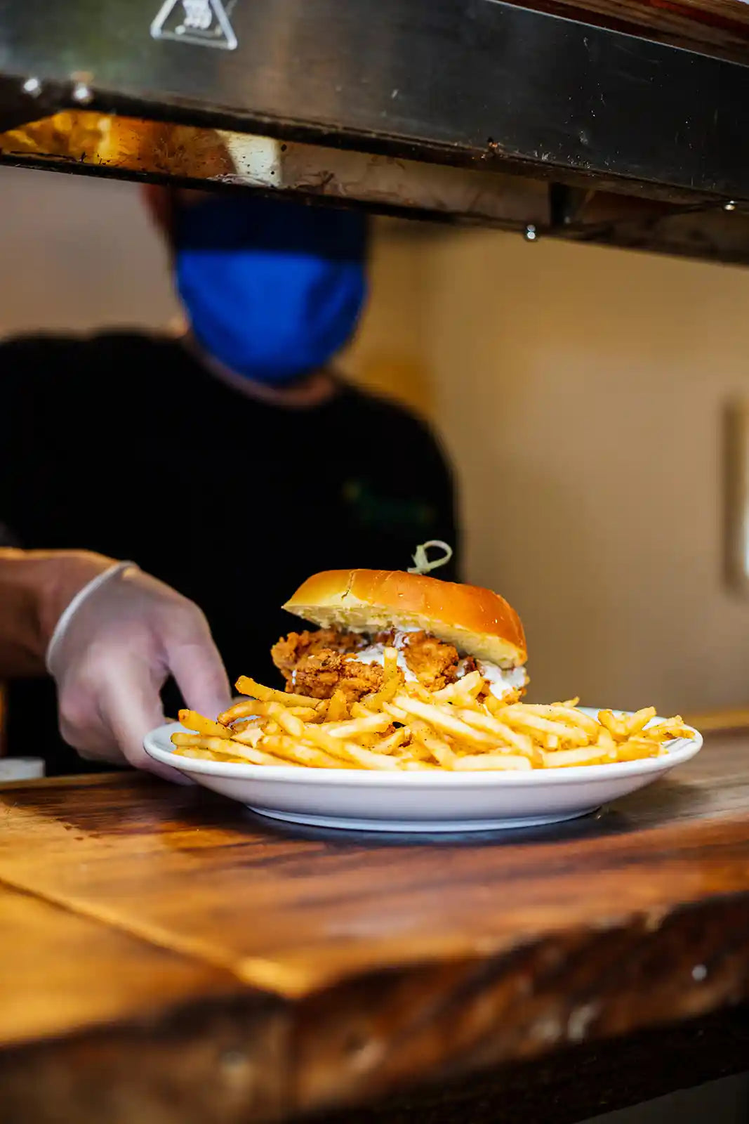 Person holding a plate with a fried chicken sandwich and fries under a heat lamp.
