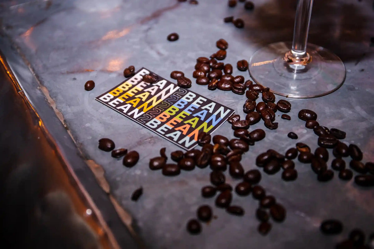 Coffee beans and 'BEAN2BEAN' stickers on a metallic surface with a glass in the background.