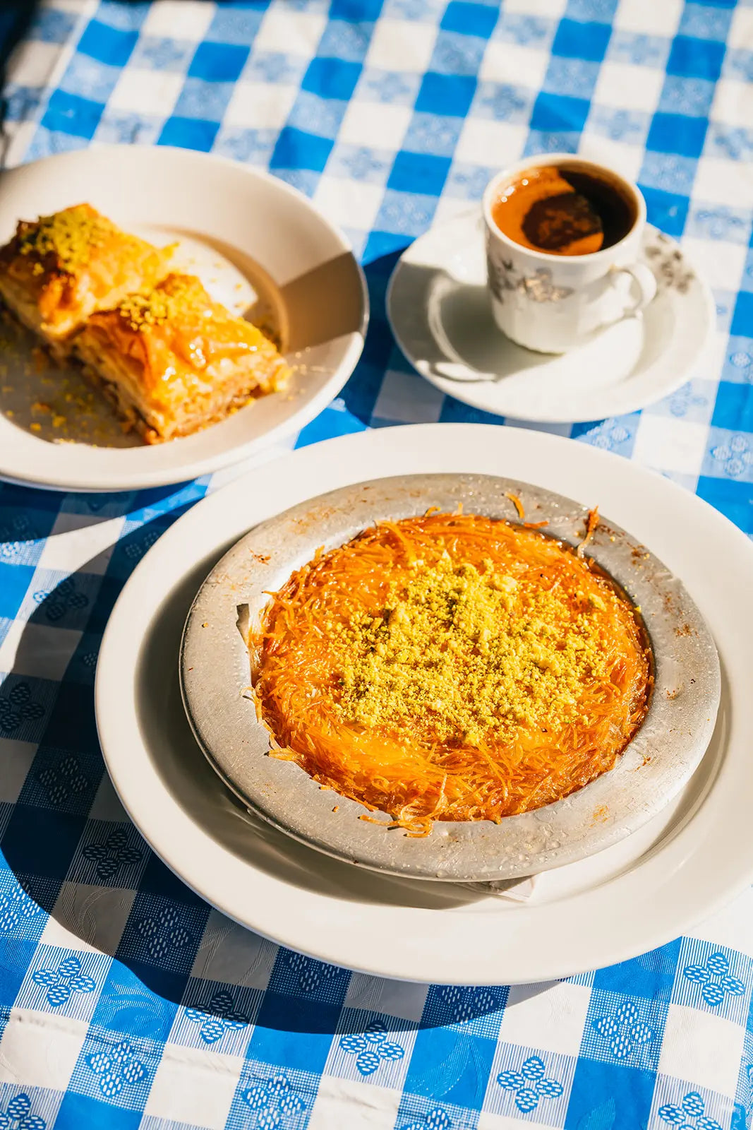Kunefe with a slice of Baklava, and a cup of Bean2Bean coffee on a blue checkered tablecloth.