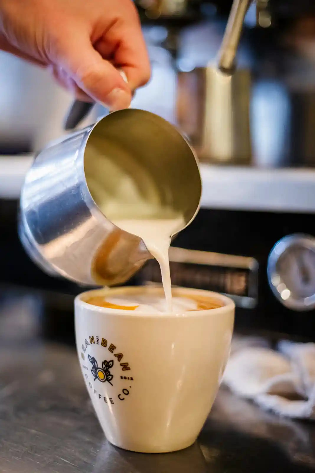 Café au lait being poured into a Bean2Bean coffee cup with a blurred coffee machine in the background