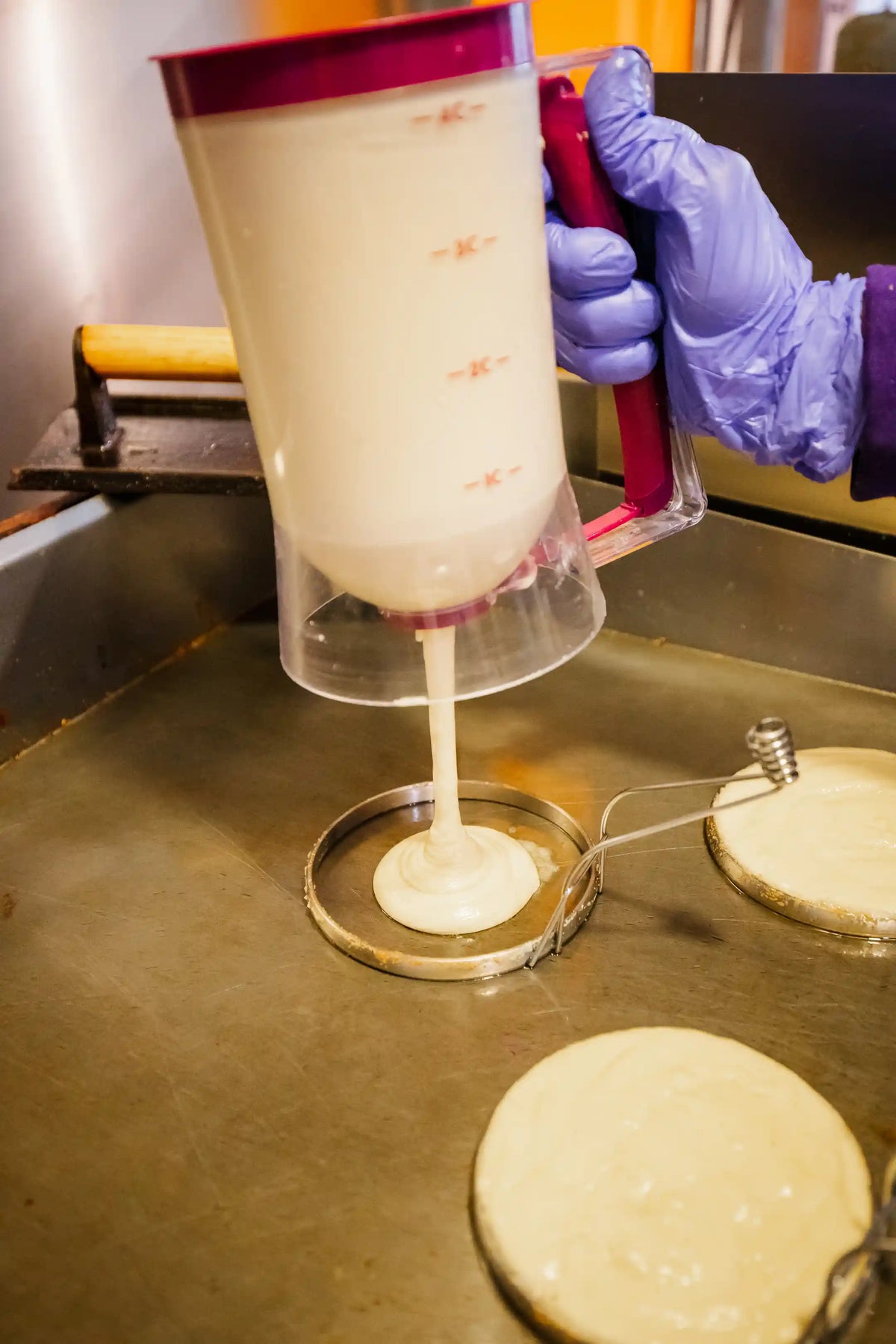 Person in purple gloves using a dispenser to pour batter onto a griddle.