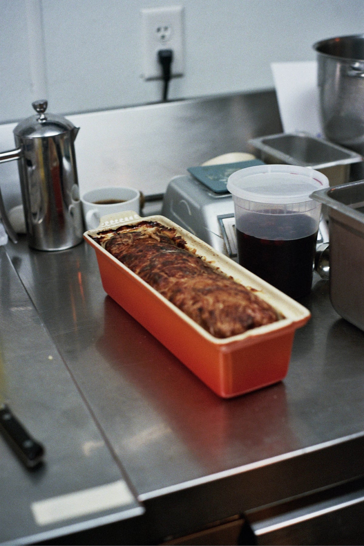 Loaf of bread or melted dish in an orange container on a metal kitchen counter with a toaster and coffee pot in the background.