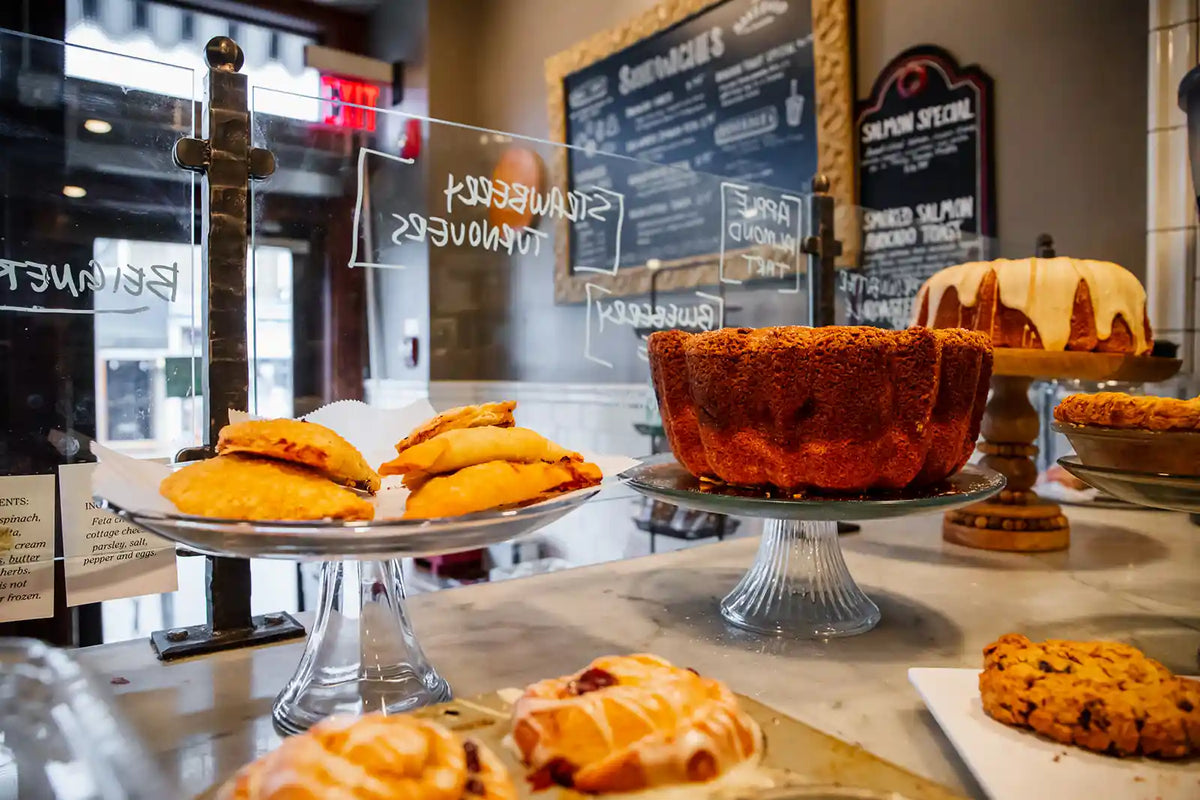 Display of pastries and cakes in a bakery setting with chalkboard menus in the background.