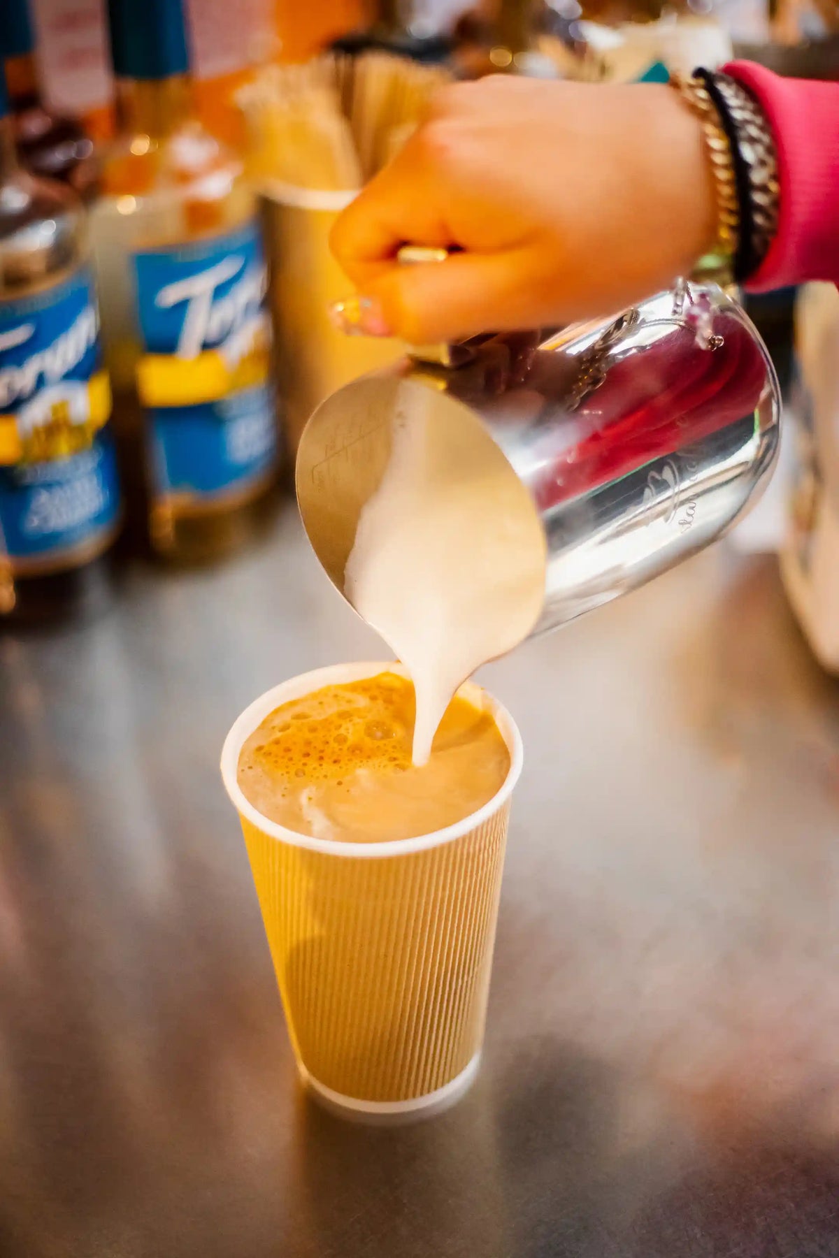Person pouring a cream into a yellow coffee cup with bottles in the background