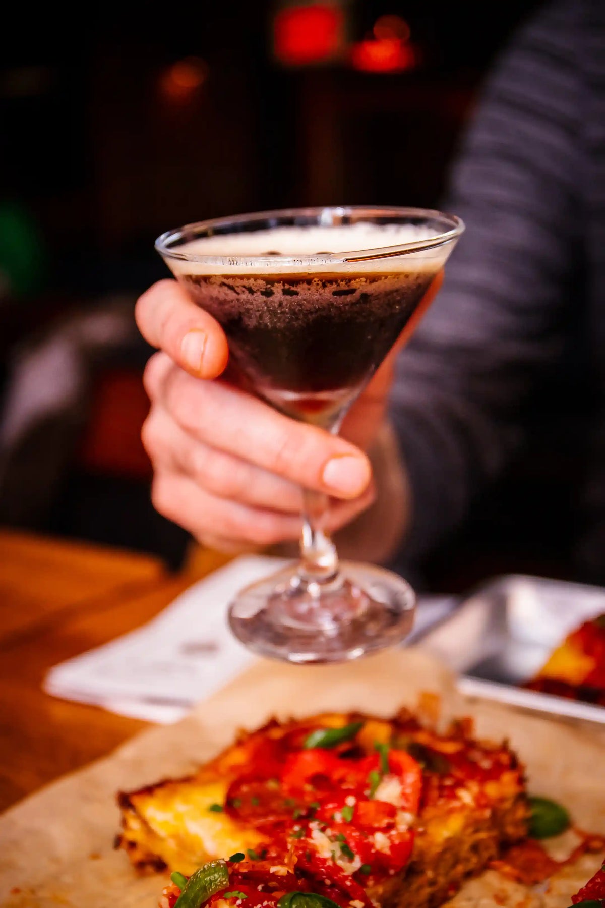 Hand holding an espresso martini cocktail glass with a dark beverage, next to a slice of pizza on a wooden table.