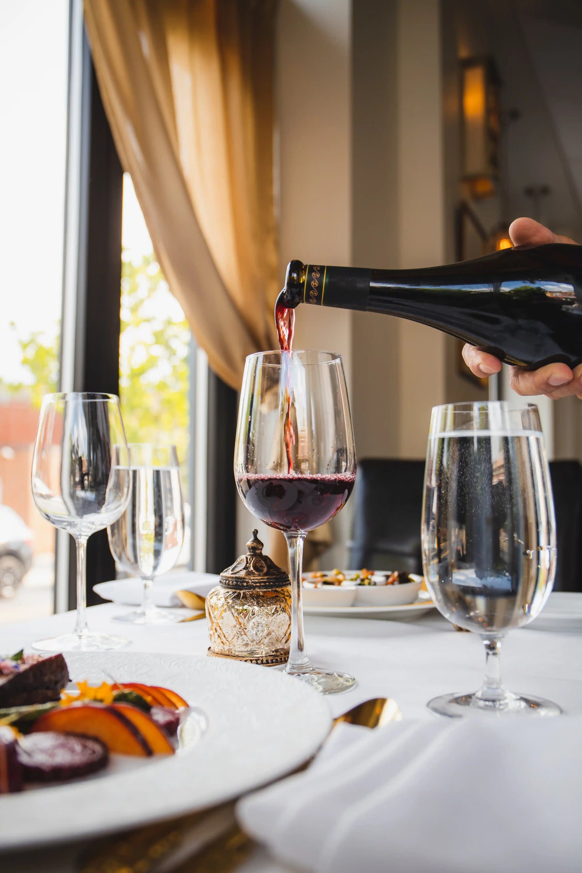 Person pouring red wine into a glass on a dining table with a scenic view.