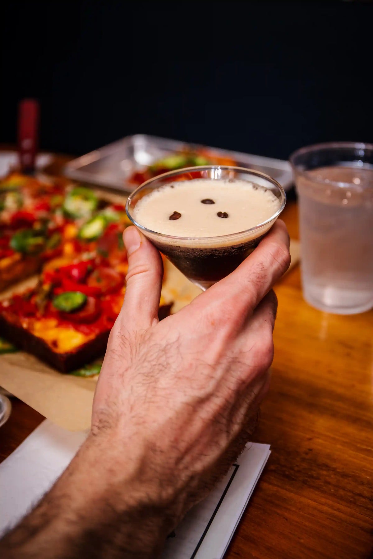 Hand holding an espresso martini with a blurred background of food and water.