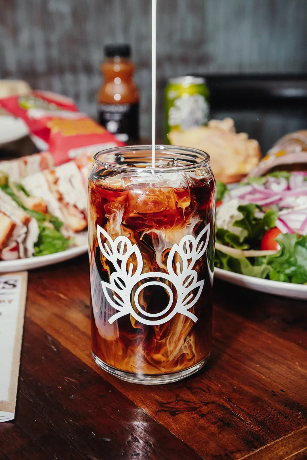 Glass of iced coffee with a Bean2Bean logo on a wooden table with food in the background