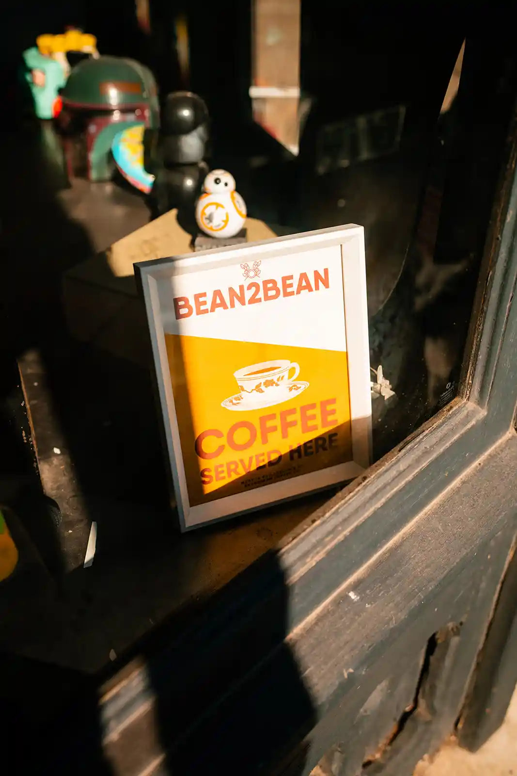 Framed sign with 'BeanzBean Coffee Served Here' on a dark surface with toy figures in the background.