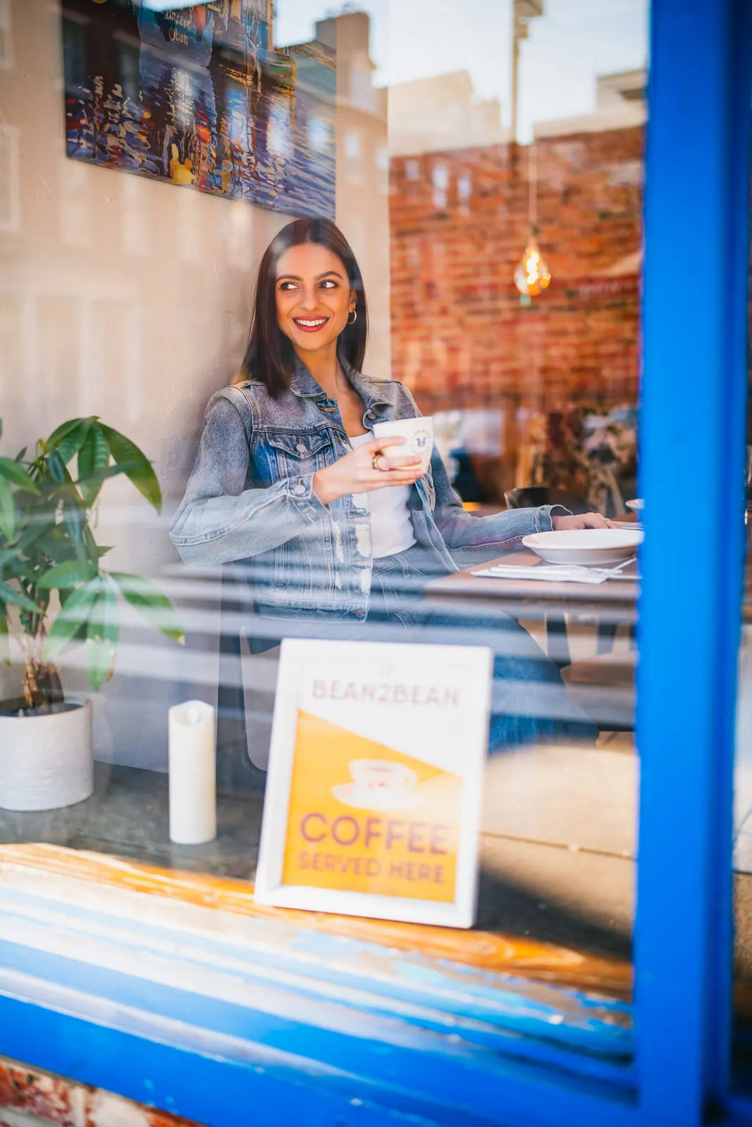 Woman sitting at a table inside Kanella Grill in a coffee shop with a 'Bean2Bean Coffee Served Here' sign.