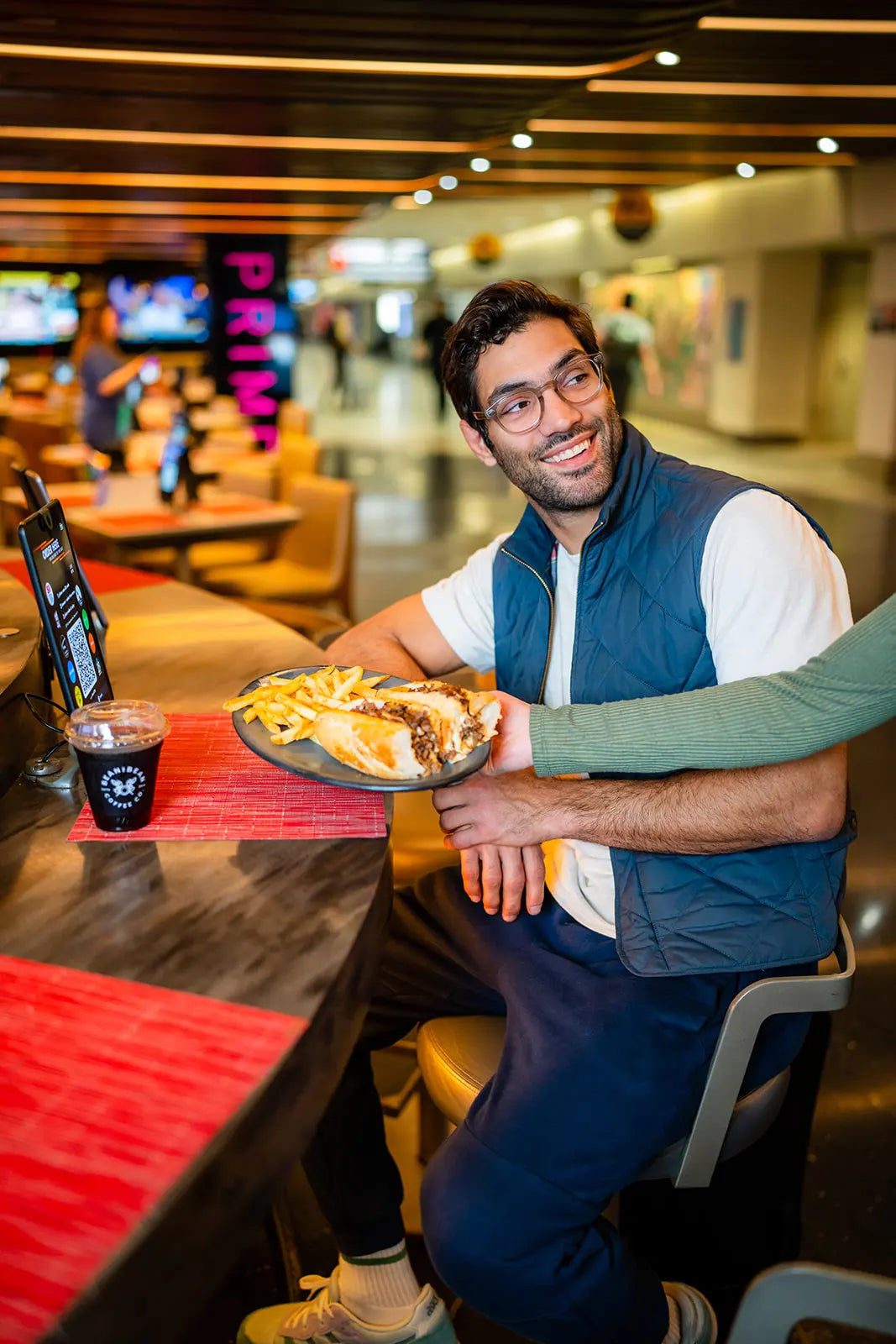 Man being served a plate with a cheesesteak and fries on it in Independence prime at a round tabletop and a Bean2Bean Cold Brew on the table
