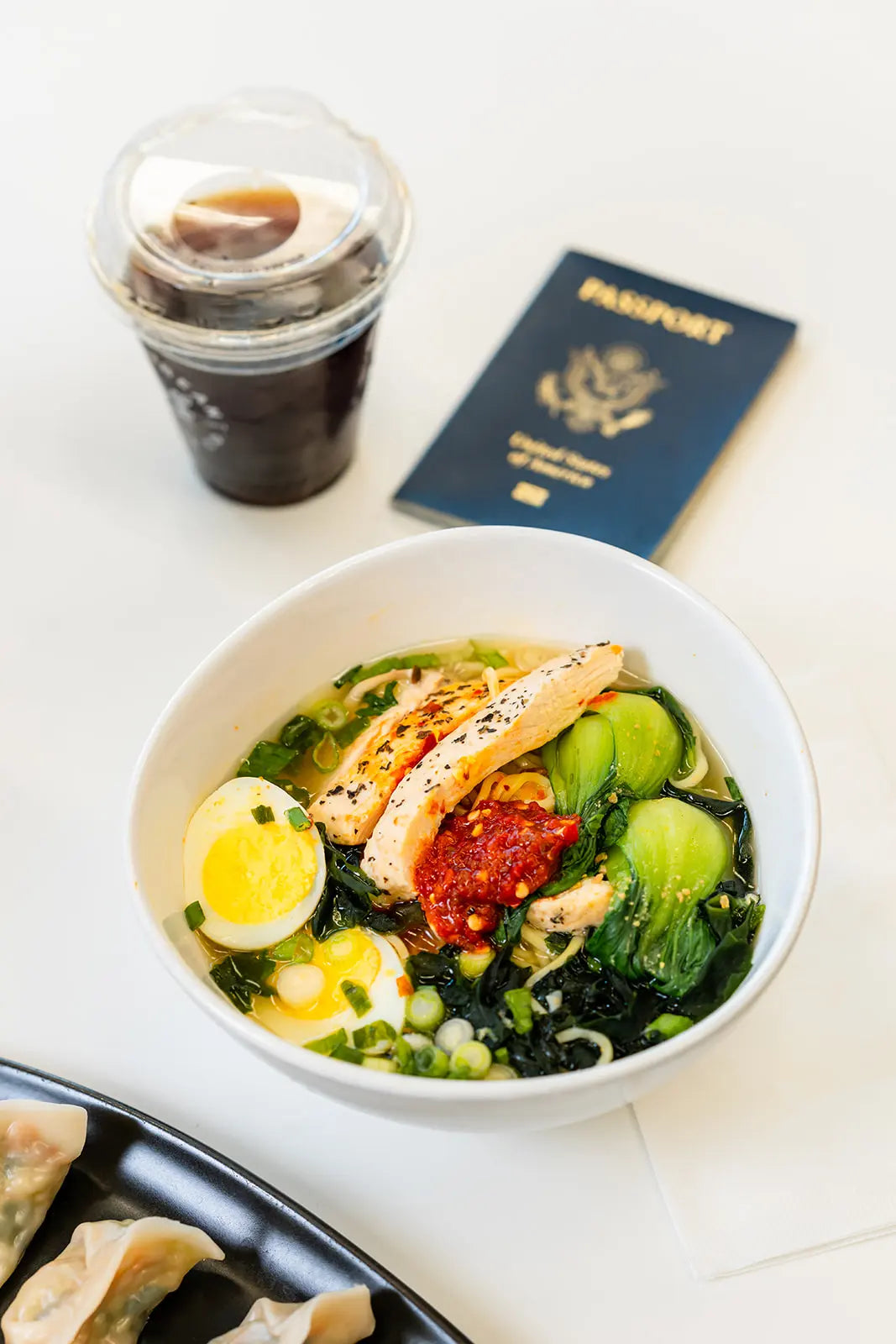Bowl of soup with vegetables and a passport on a white surface near a Bean2Bean Cold Brew