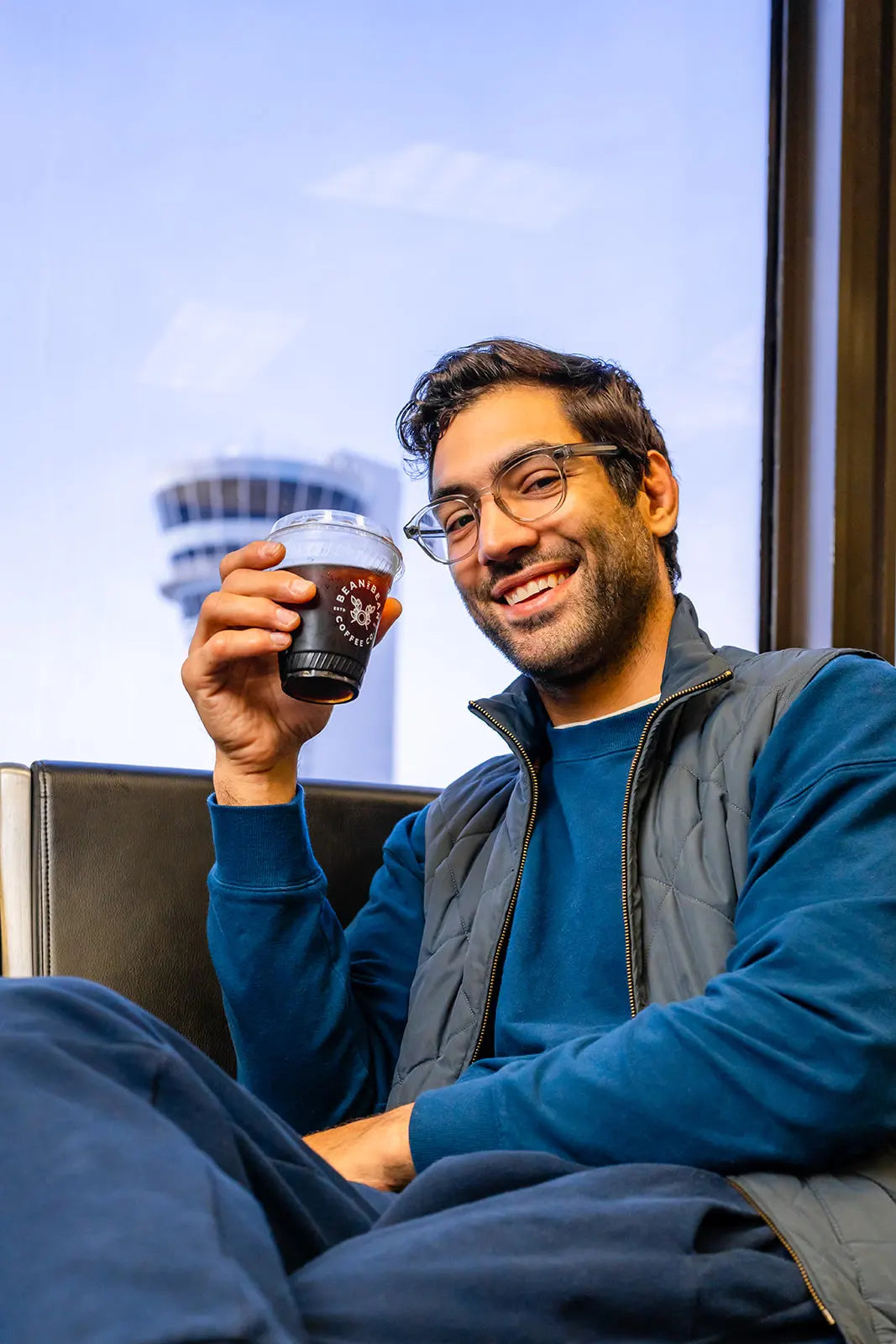 Man sitting by a window at PHL Airport holding a Bean2Bean Cold Brew, with the control tower in the background