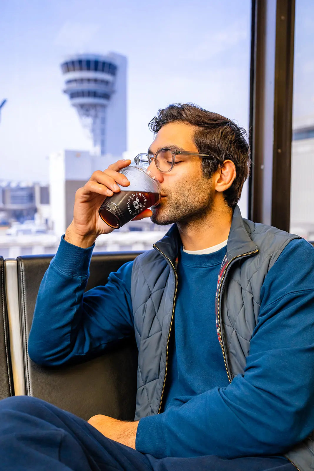 Man sitting by a window at PHL Airport drinking a Bean2Bean Cold Brew, with the control tower in the background