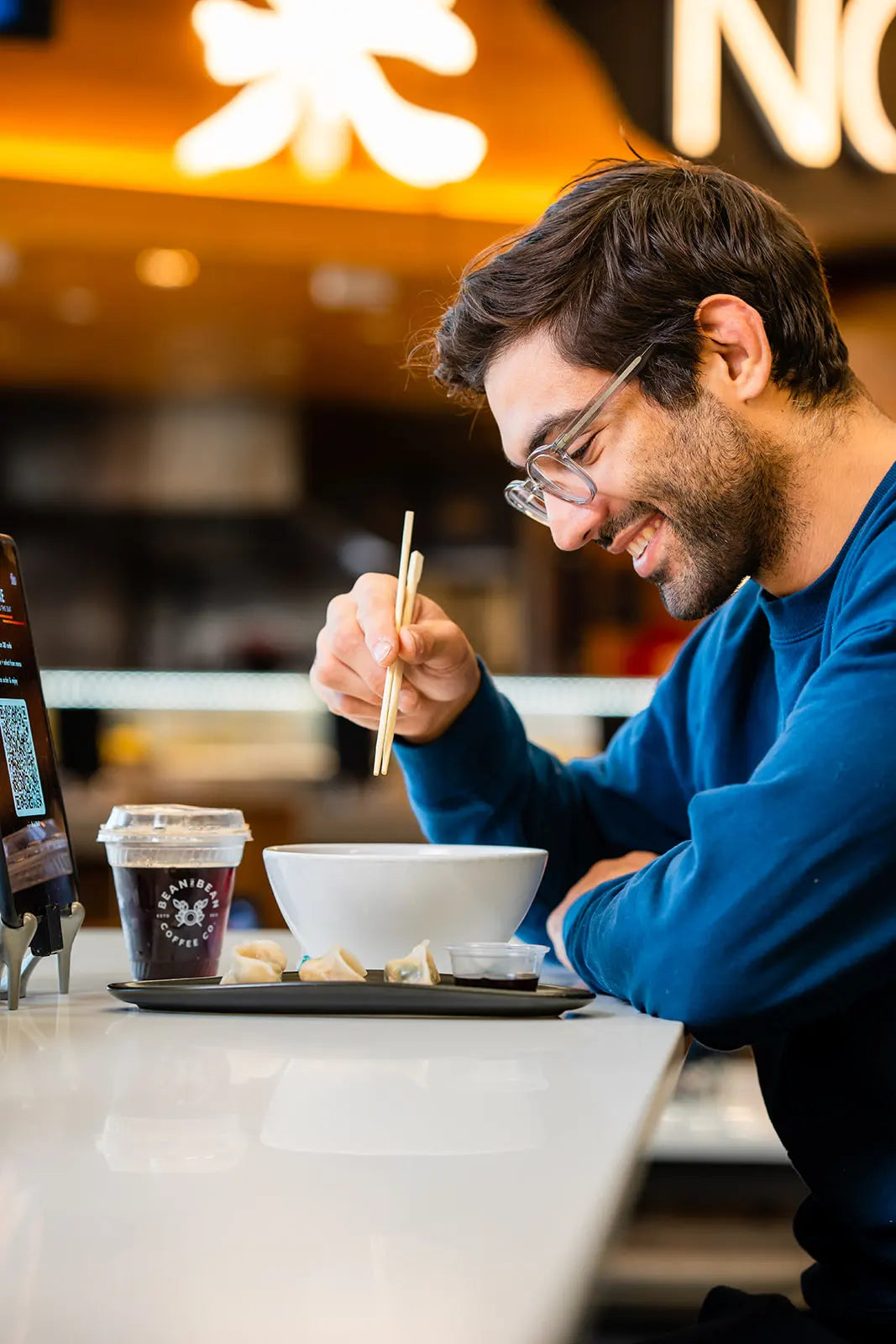 Man eating out of a bowl with chopsticks and a Bean2Bean cold Brew in front of him at a Noobar
