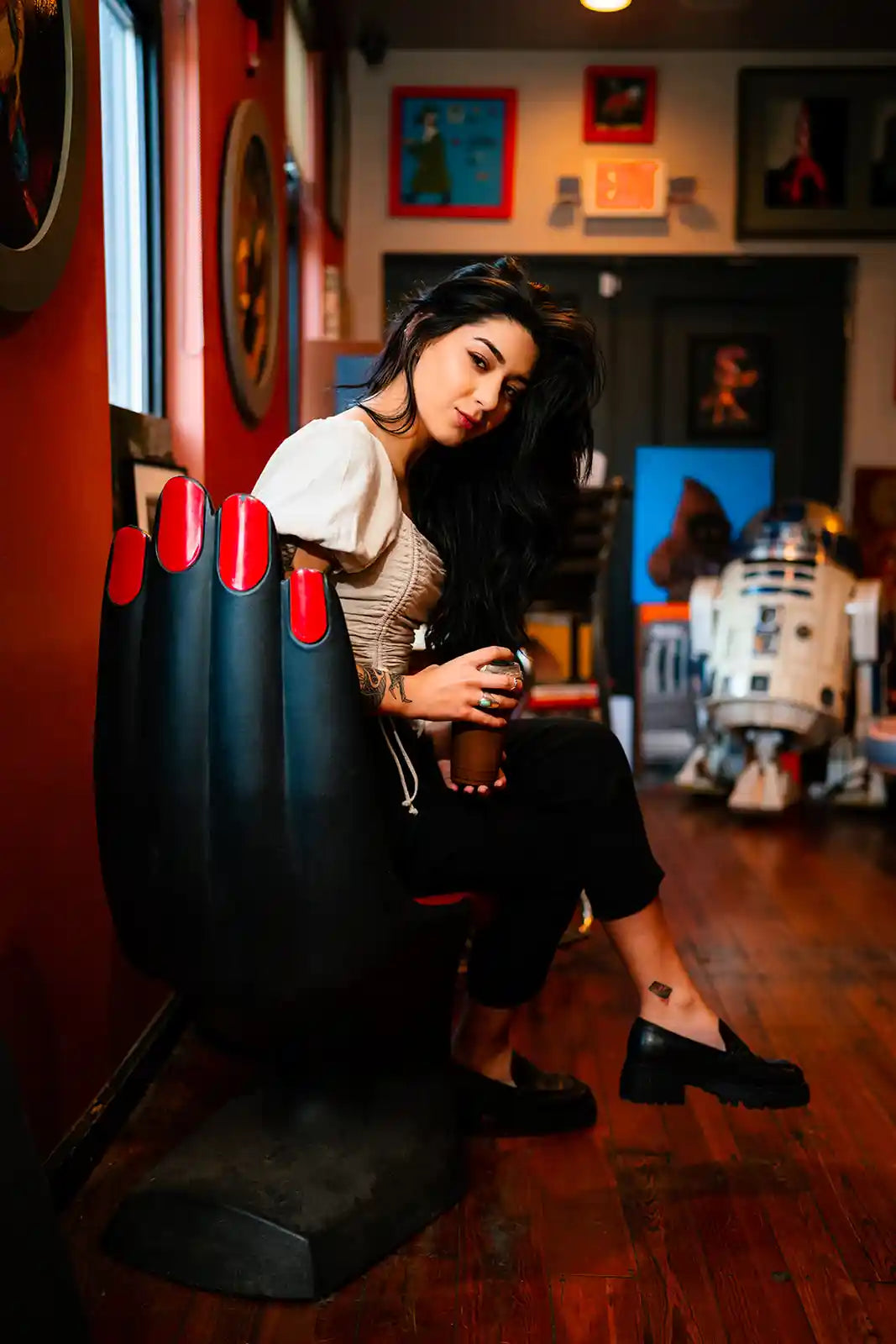 Woman sitting on a large black shoe in a room with various decorations and a robot figure.