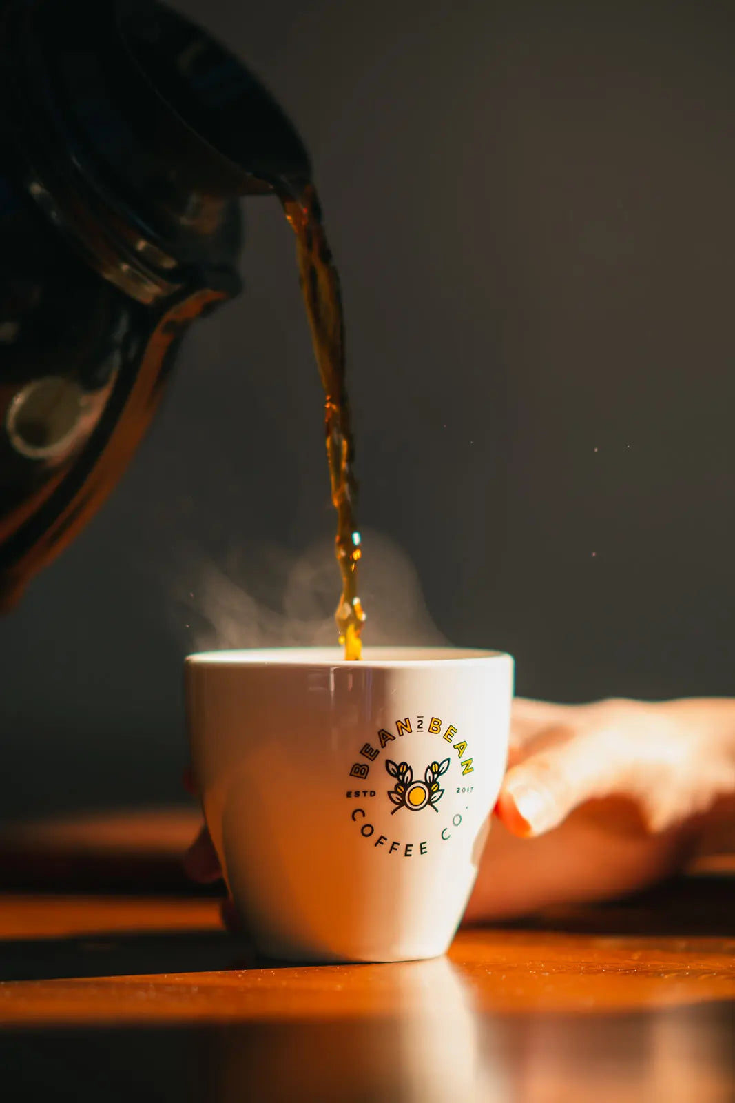 Bean2Bean Coffee being poured into a white Bean2Bean cup with a visible logo on a wooden surface.