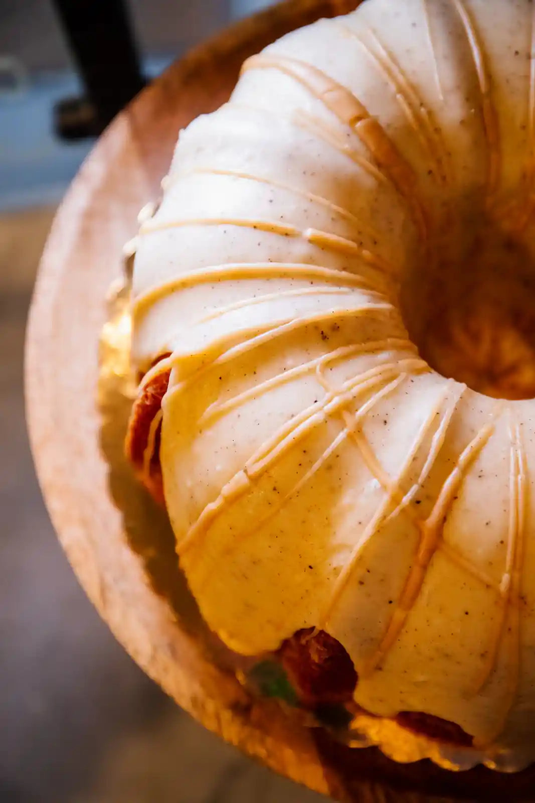 Close-up of a bundt cake with a white frosting and a drizzle of orange sauce on a wooden stand.