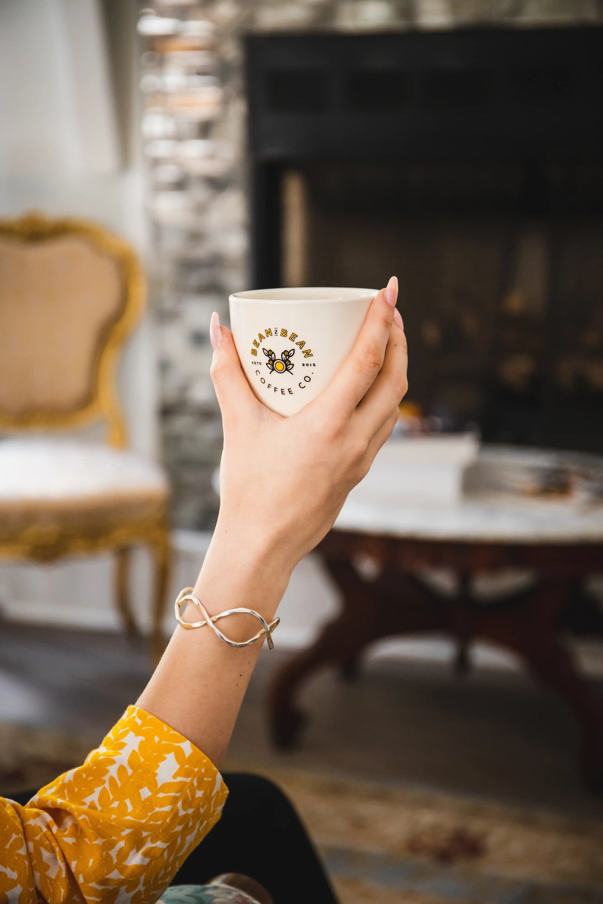 Hand holding a Bean2Bean coffee cup with a brand logo in a cozy indoor setting.