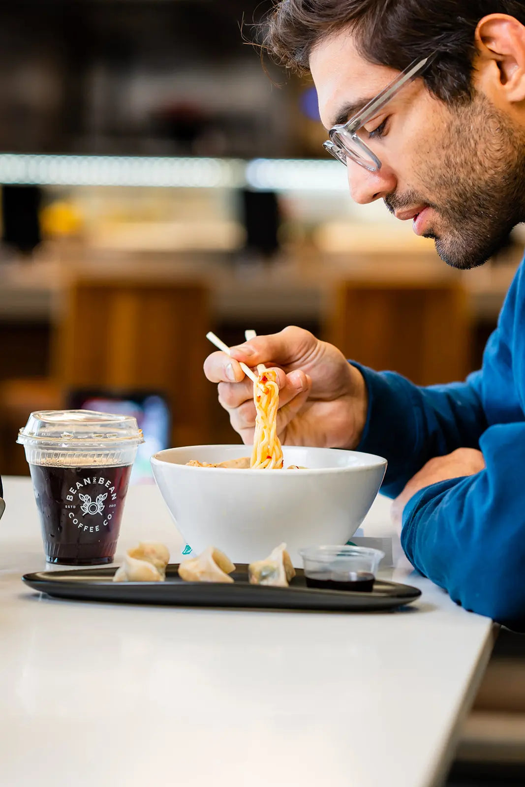 Man eating noodles out of a bowl with chopsticks and a Bean2Bean cold Brew in front of him at a Noobar