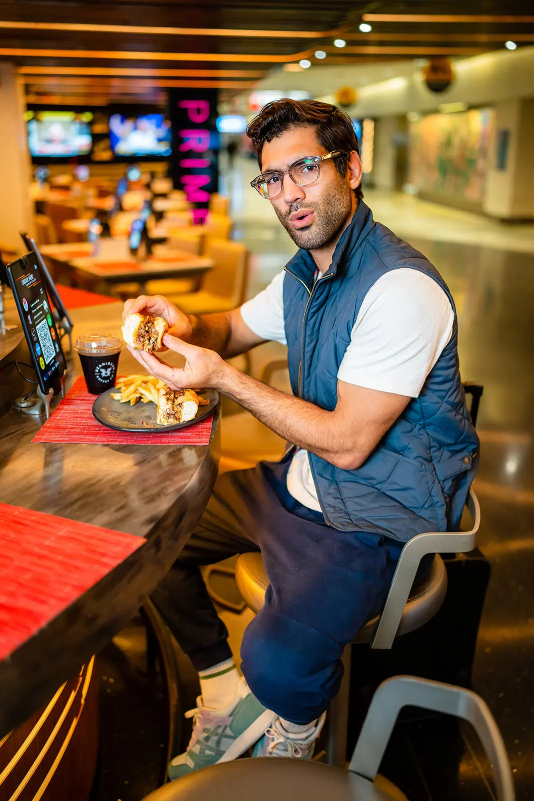 Man eating a cheesesteak sandwich at a Independence Prime restaurant table with neon signs in the background