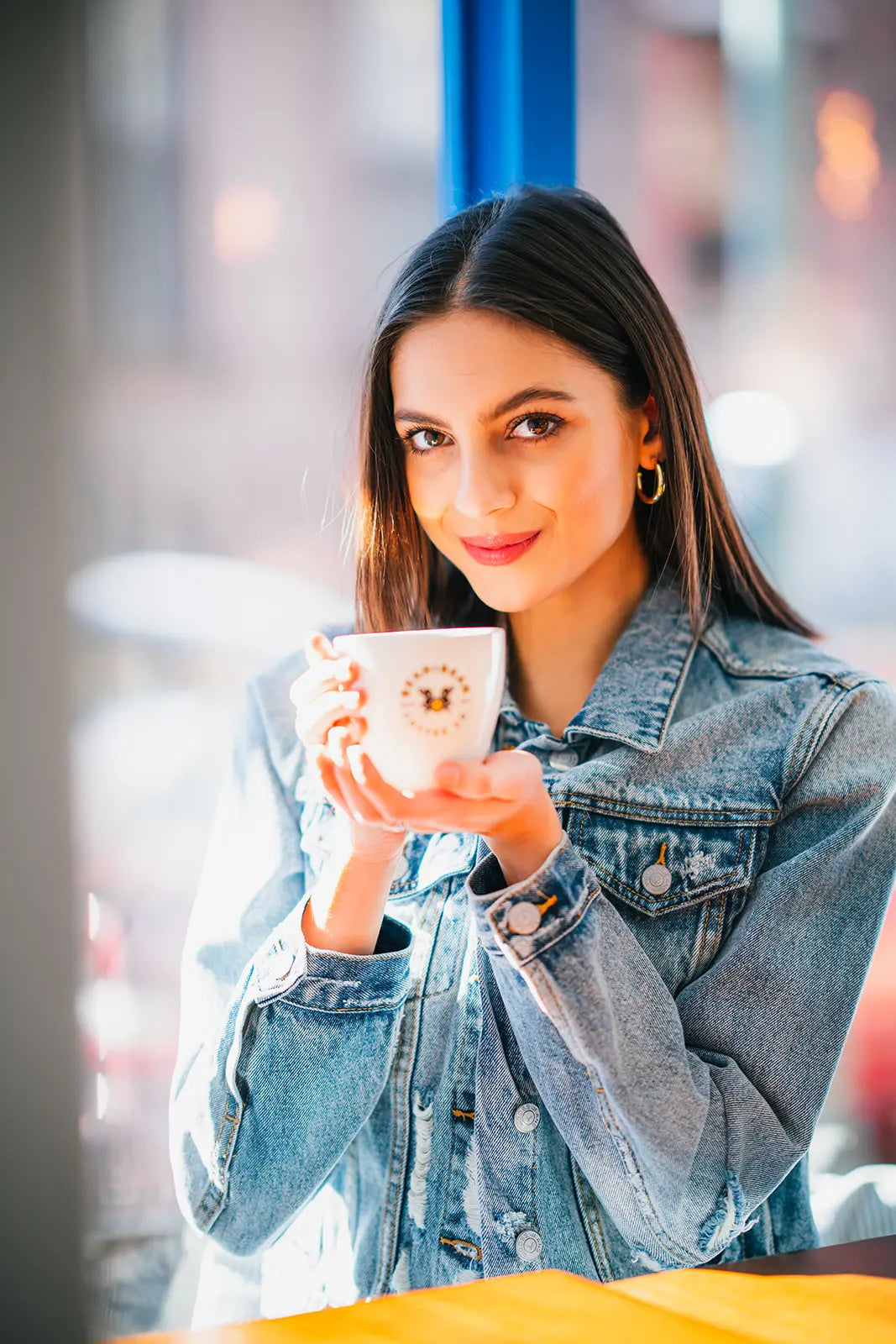Woman holding a white Bean2Bean mug with a decorative design, wearing a denim jacket indoors.